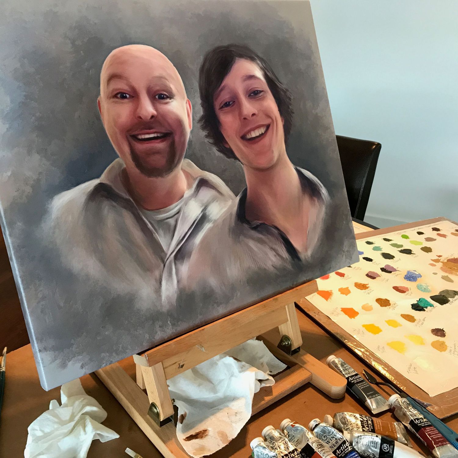 Oil painting in progress showing a memorial portrait of father and son with artist's palette visible.