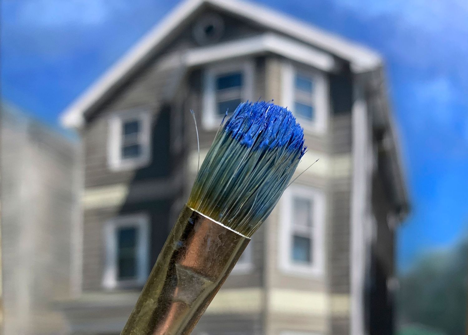 A blue-tipped paintbrush against a an out of focus house painting in the background.