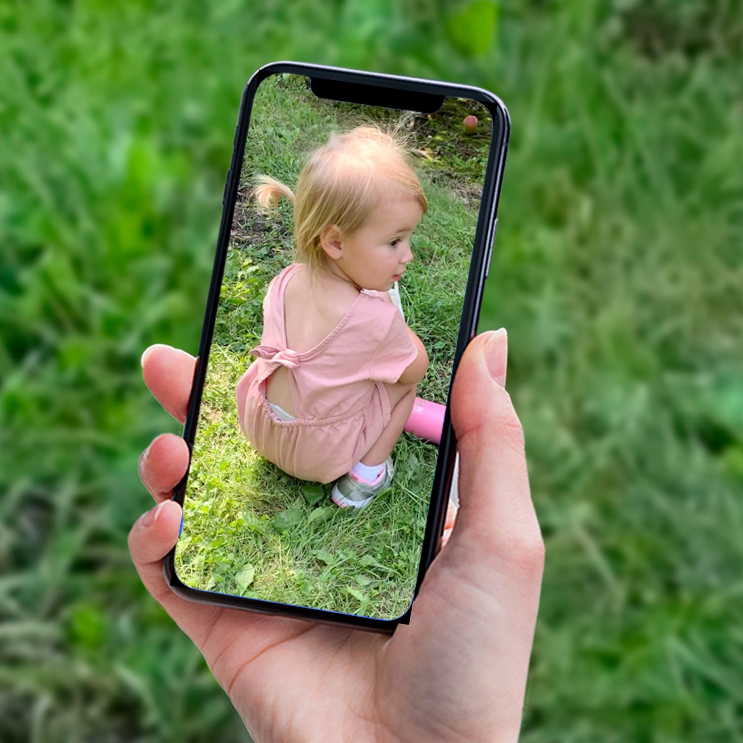 Smartphone displaying photo of a toddler in pink clothing sitting in green grass. Source photo for a painting commission.