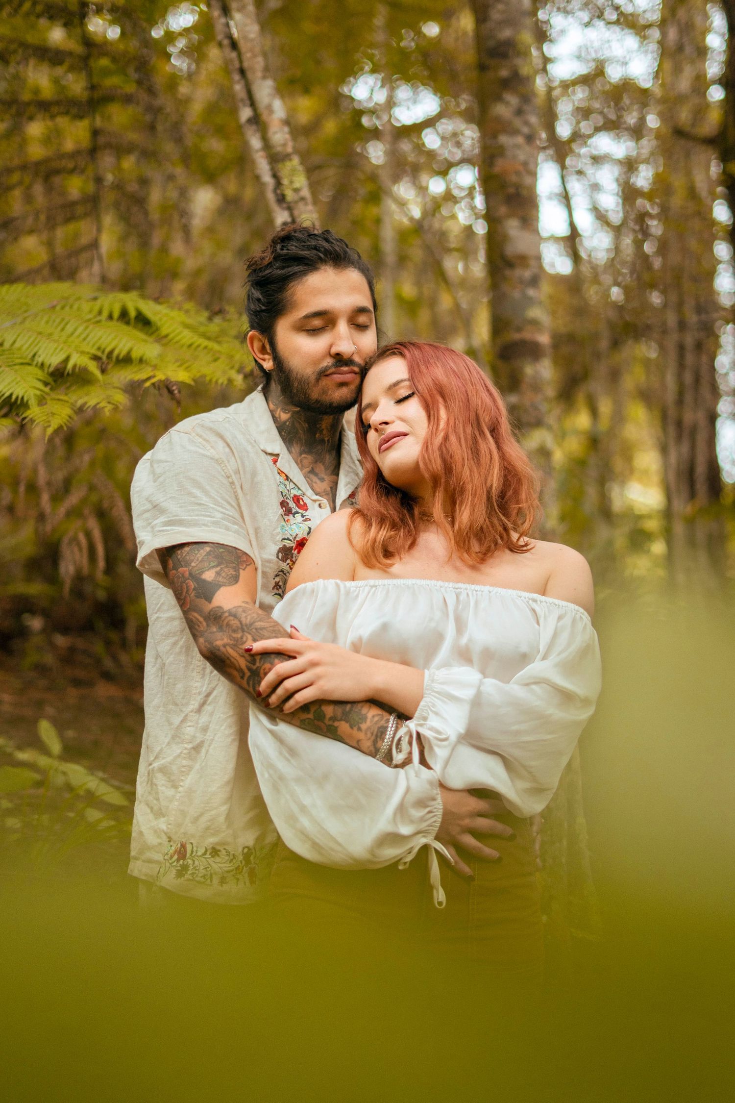 Couple embracing in forest, eyes closed, sharing a peaceful, romantic moment surrounded by lush greenery and soft light.