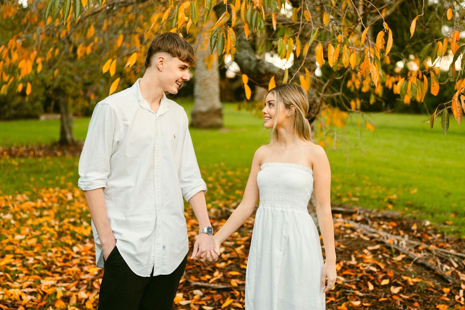Young couple holding hands and smiling under autumn trees in a park, dressed in white and surrounded by fallen golden leaves.