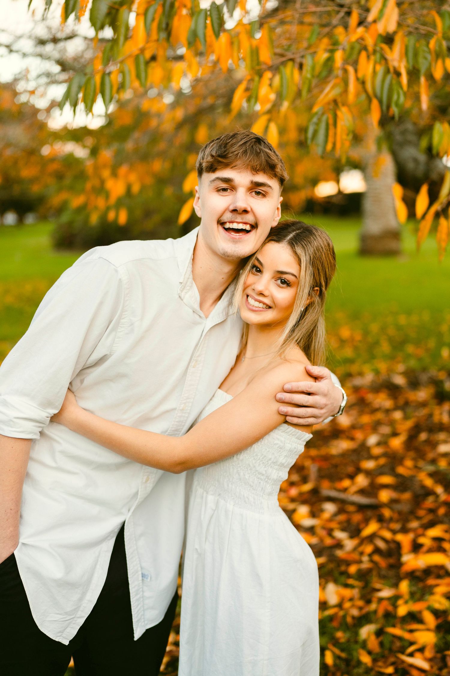 Young couple smiling and hugging under autumn leaves in a park, wearing white outfits and enjoying a golden sunset moment.