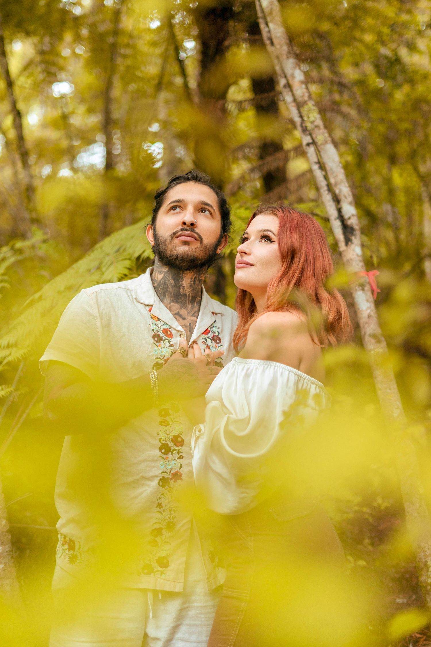 Couple standing in lush forest, gazing upward with serene expressions, surrounded by golden foliage and natural light.