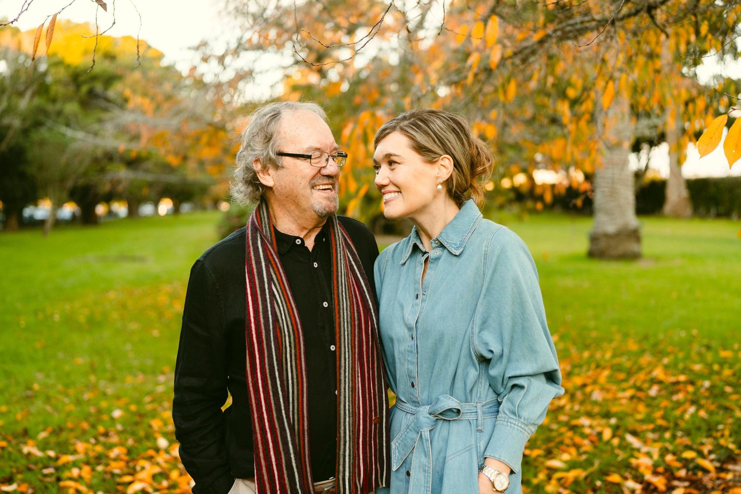 Smiling man and woman standing together in a park during autumn, surrounded by colorful leaves and trees.