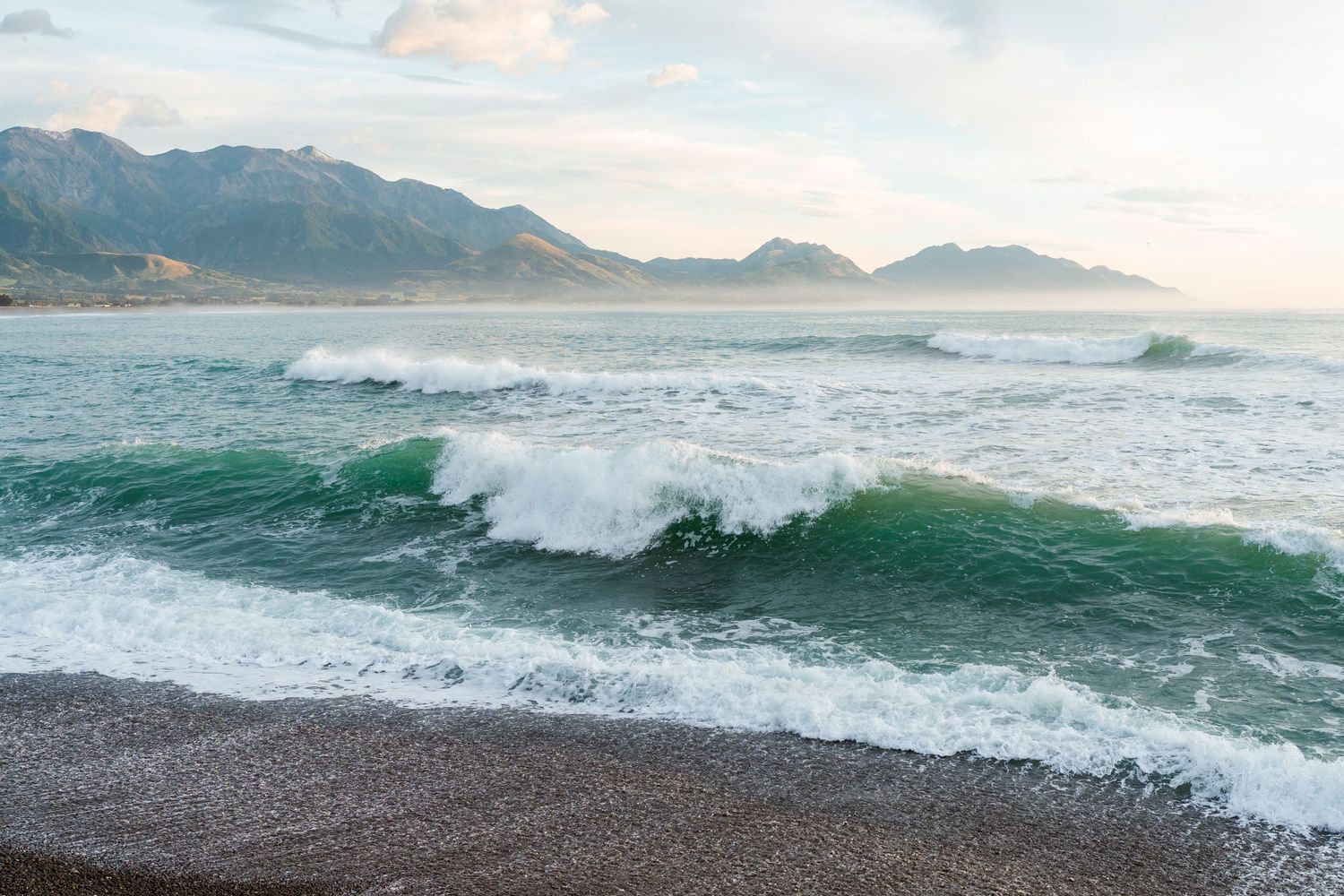 Waves crashing on Kaikōura beach with mountain ranges in the background under a soft pastel sky at sunset.
