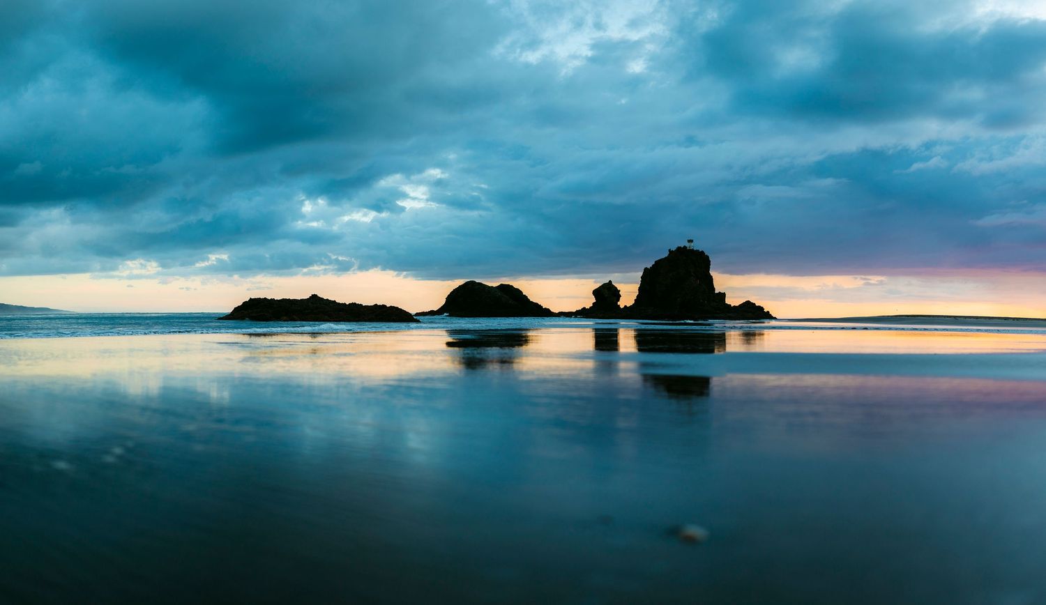 Moody coastal seascape at sunset with dramatic clouds, rugged rock formations, and serene ocean reflections at Whatipu Beach.