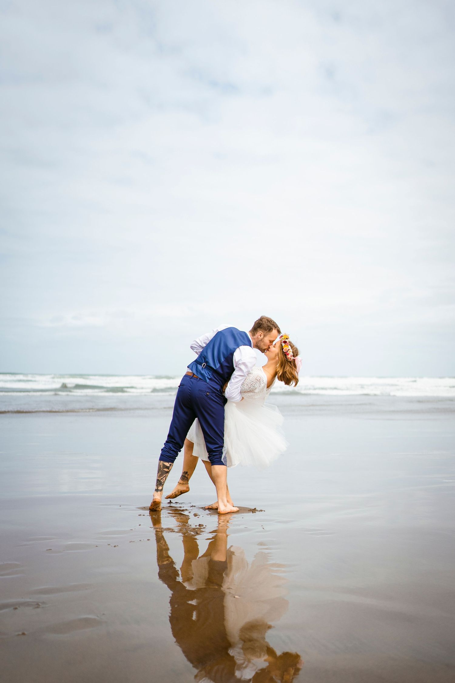 Groom dips and kisses bride on a reflective beach at low tide, barefoot and joyful under a cloudy sky. Romantic seaside wedding moment.