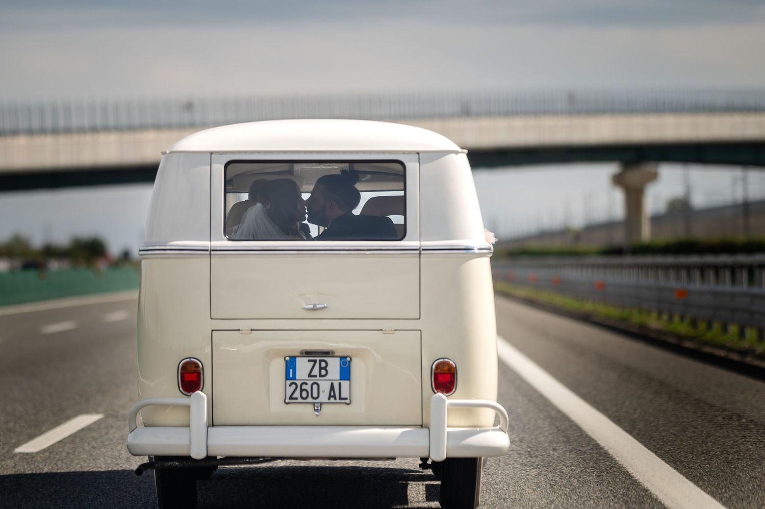 Classico furgone VW vintage bianco che guida in autostrada con ponte sullo sfondo e sposi.