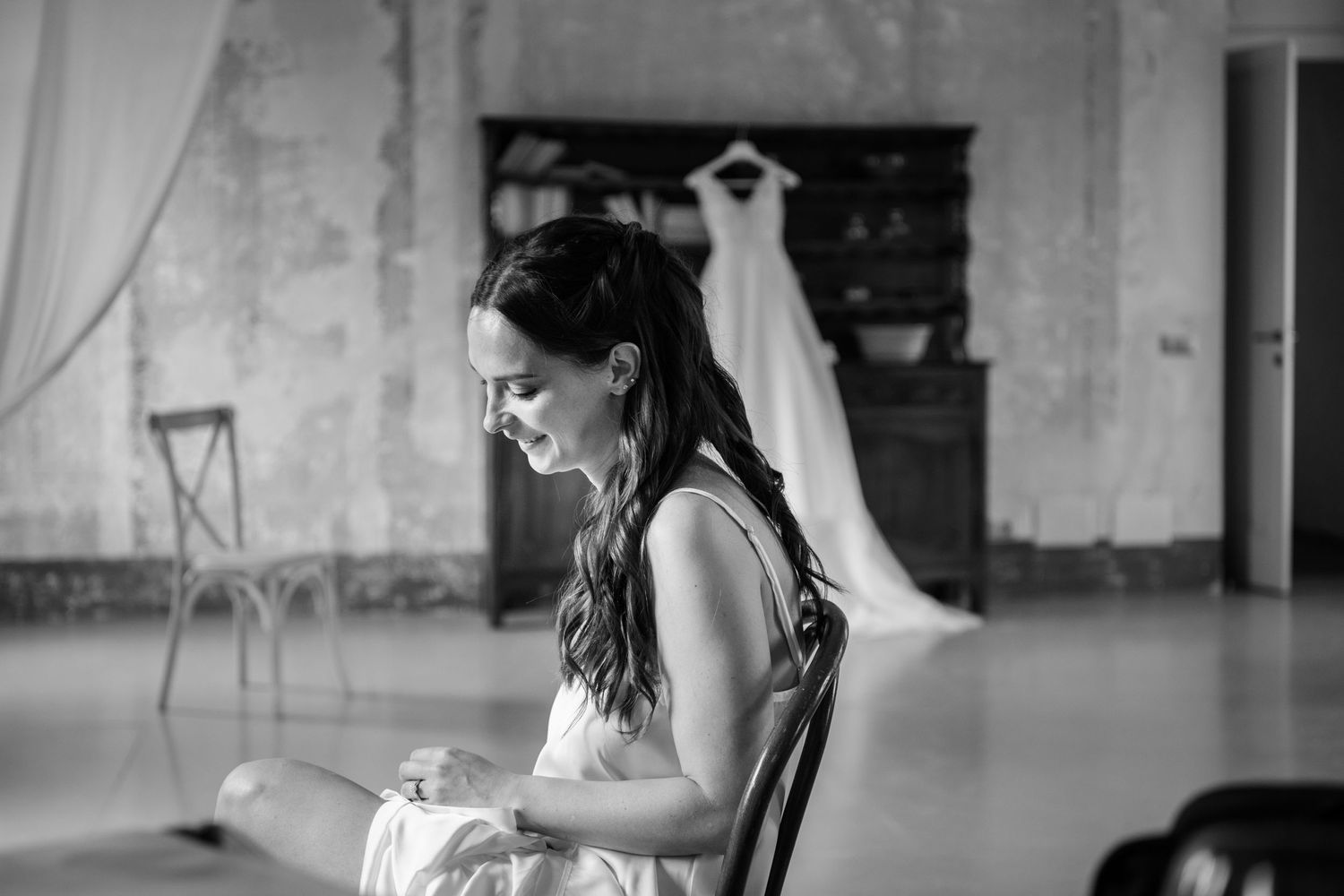 Una foto in bianco e nero mostra una sposa in abito formale seduta su una sedia in una stanza con un abito da sposa sullo sfondo.