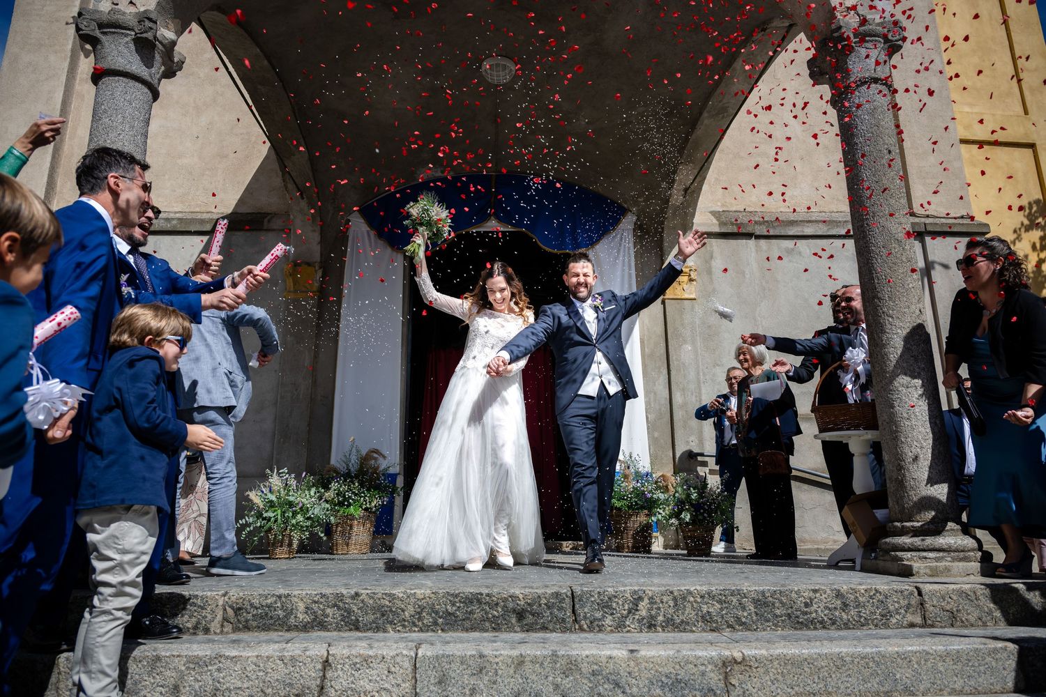 Coppia di sposi festeggia sotto l'arco con gli ospiti che lanciano coriandoli rossi e riso.