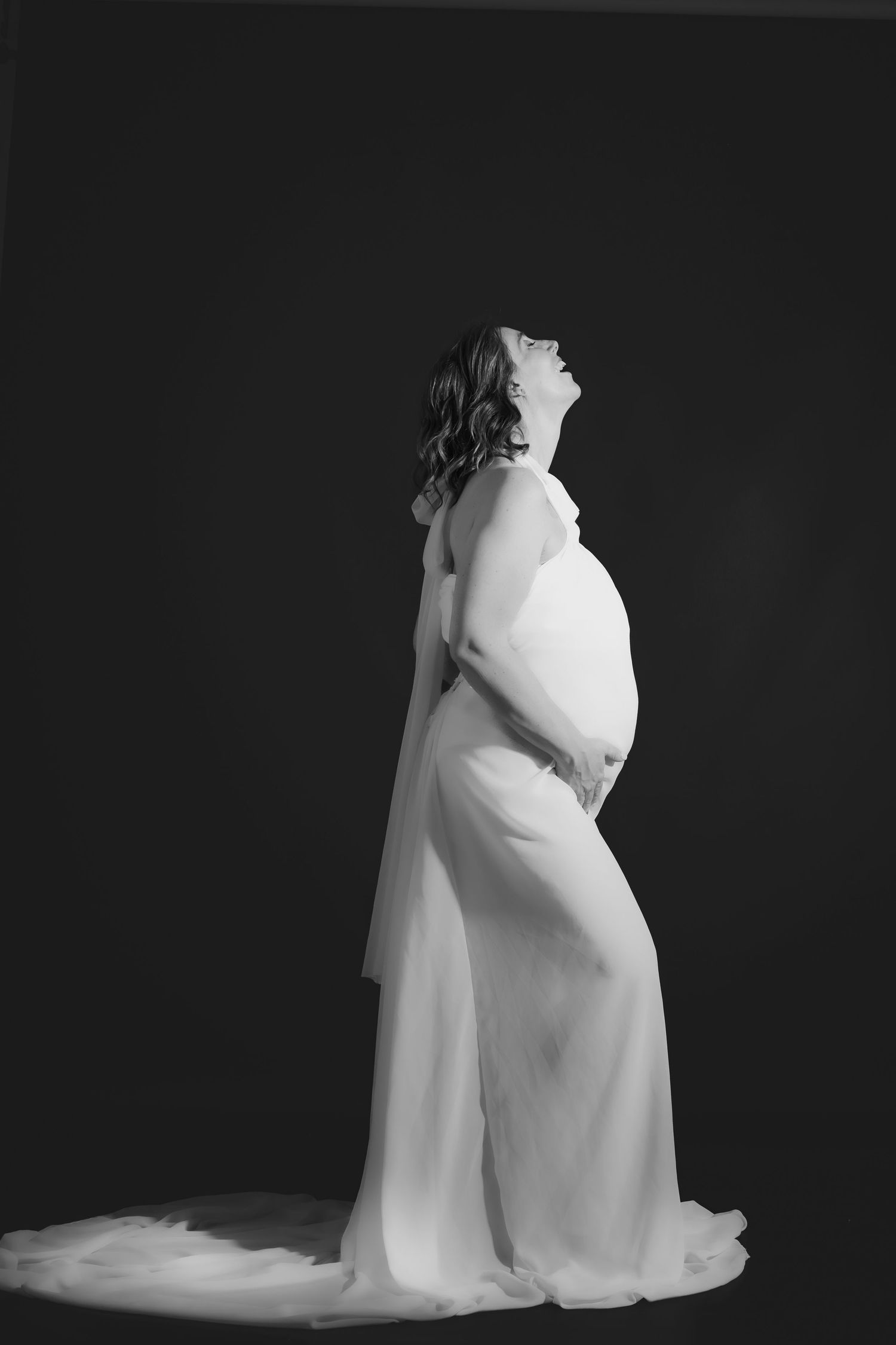 Artistic black and white maternity portrait showing a woman in flowing white gown looking upward against dark background.