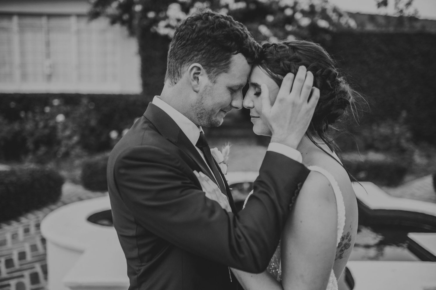 Intimate black and white wedding portrait of couple touching foreheads tenderly in outdoor garden setting.