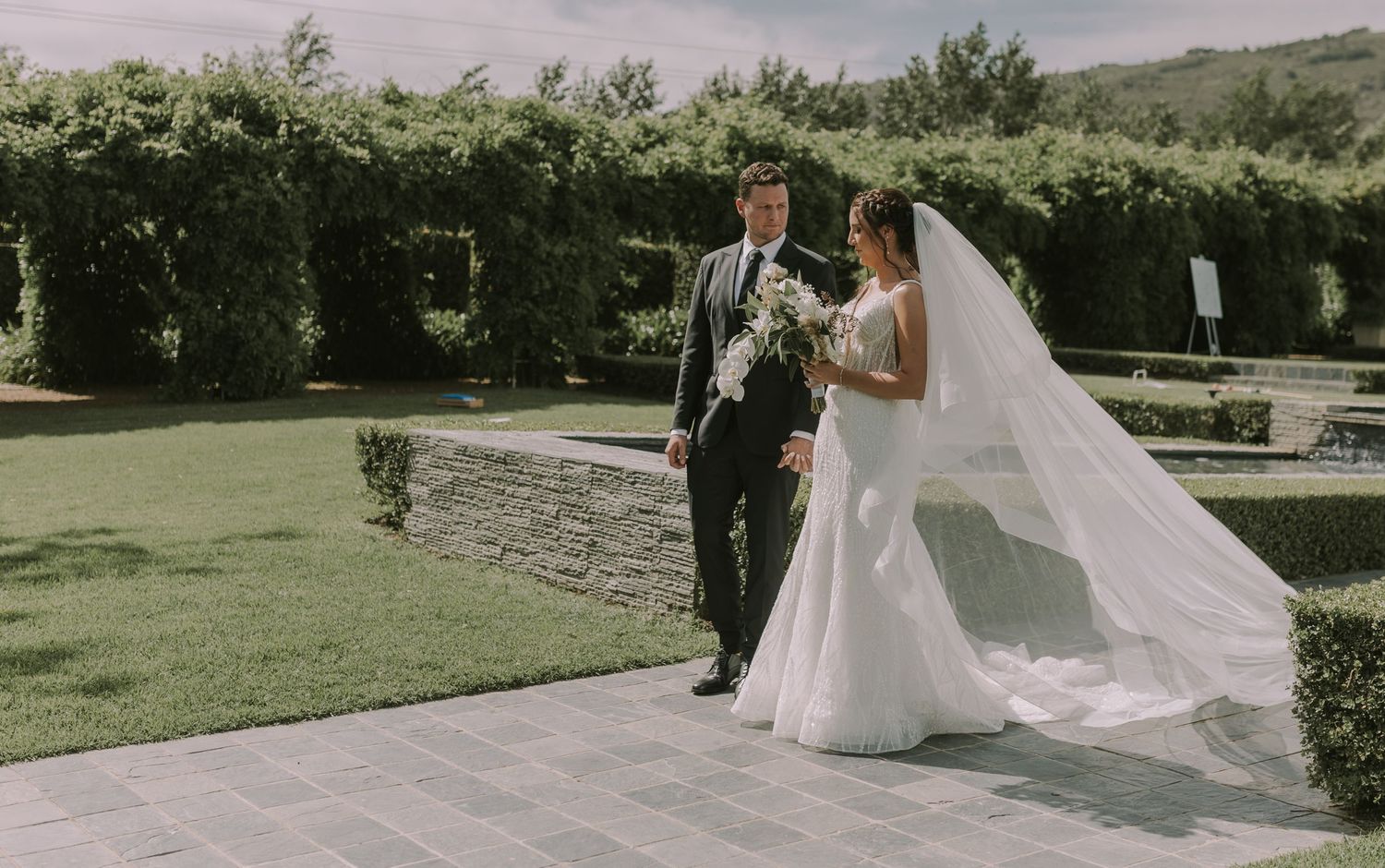 Bride and groom walk together through a lush garden venue on their wedding day.