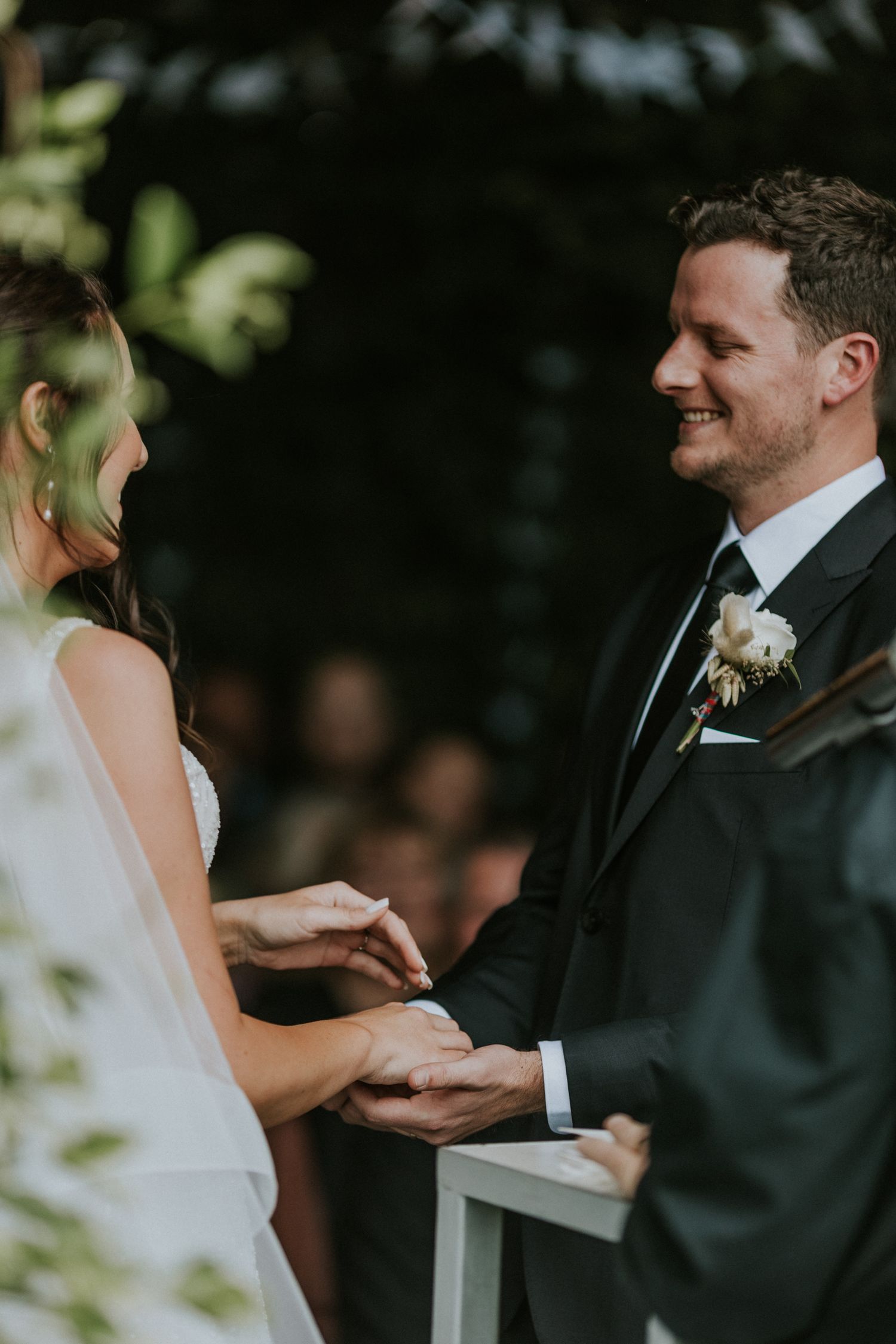 Joyful groom smiling at bride while exchanging rings during outdoor wedding ceremony.