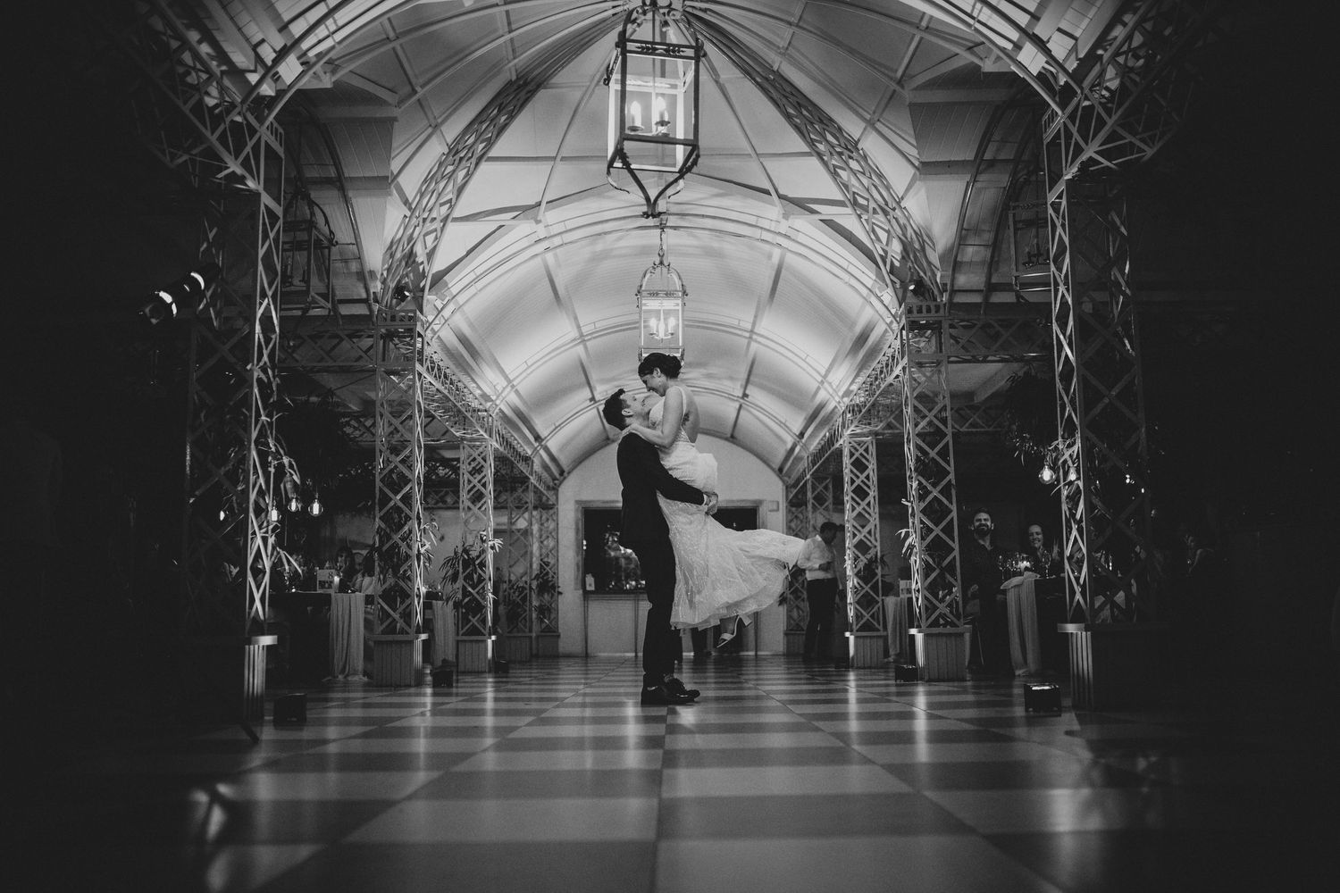 Bride and groom share romantic dip kiss in ornate arched hallway with chandeliers.