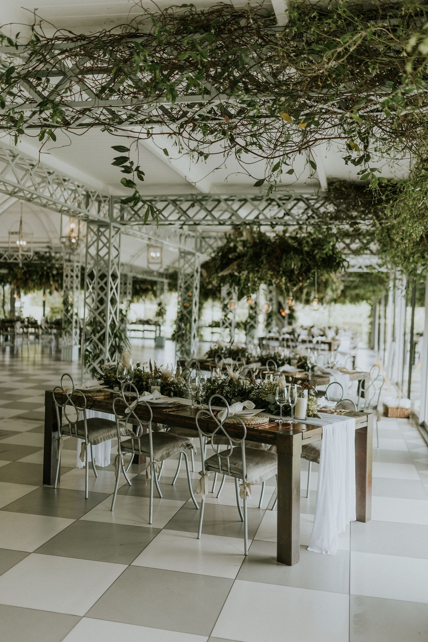 Elegant wedding reception venue with greenery ceiling installation, white lattice columns, and styled dining tables on checkered floors.