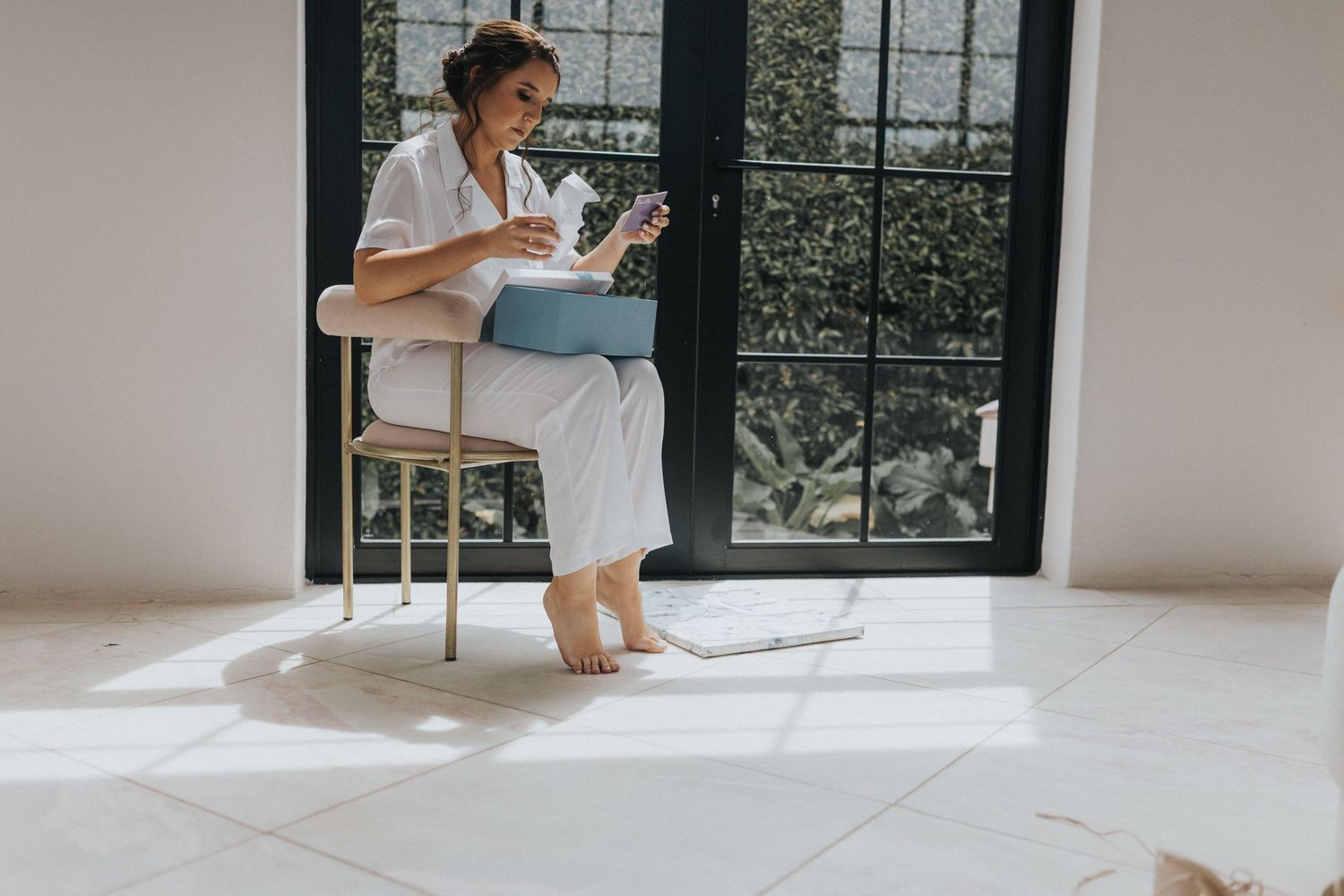 Bride in white robe sits barefoot on chair reading near large glass doors.