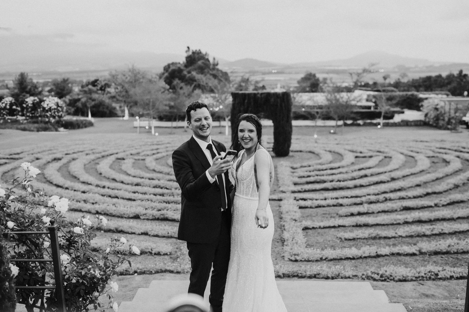 Black and white photo of smiling bride and groom standing before a circular garden labyrinth on their wedding day.