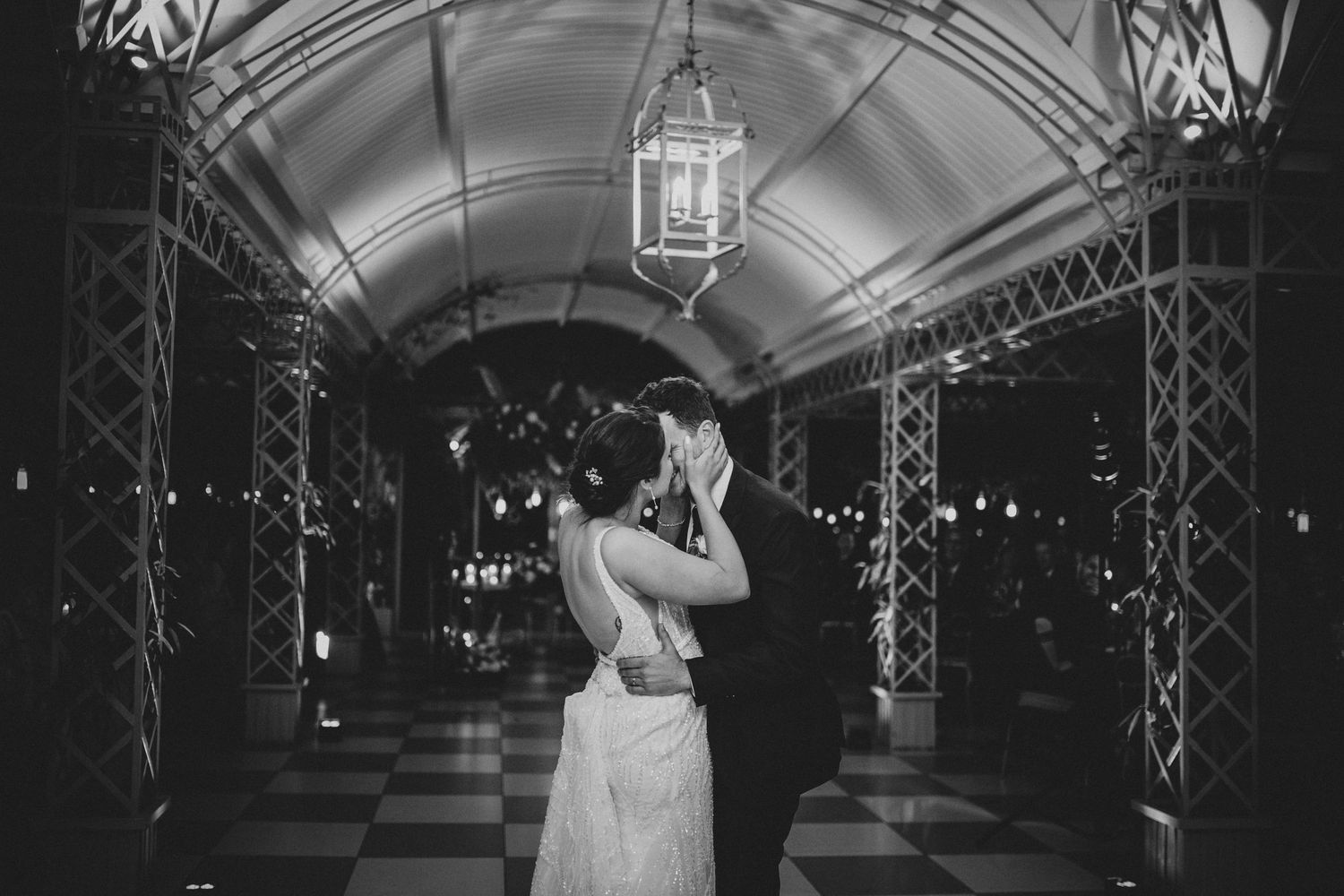 Bride and groom share intimate embrace in elegant arched corridor with checkered floors.