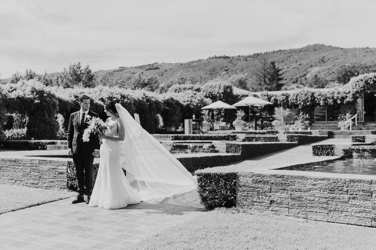 Black and white photo of bride and groom walking through elegant garden venue.