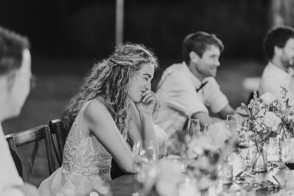 Emotional bride listens to wedding speech at outdoor reception dinner table.