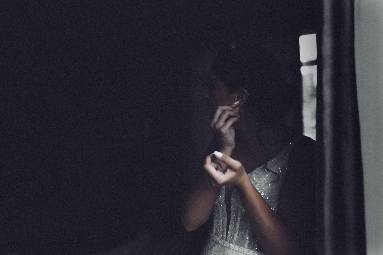Emotional bride in silver dress standing by dark window in contemplative wedding moment.