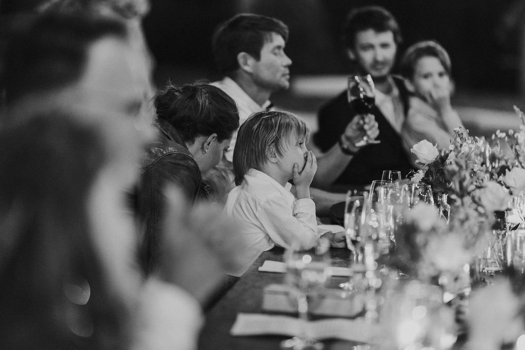 Young child covers mouth while seated at an elegant wedding reception dinner table in black and white.