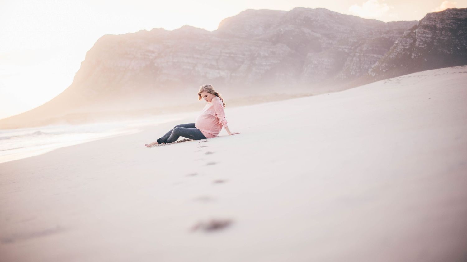 Young woman sitting alone on sandy beach with mountain backdrop and footprints in sand.