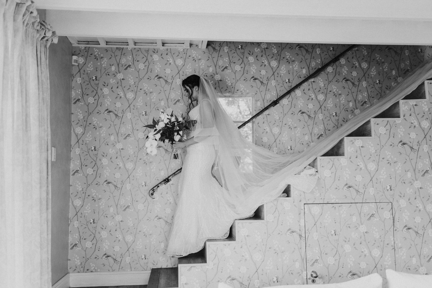 Black and white image of bride in fitted gown carrying bouquet down staircase with veil.