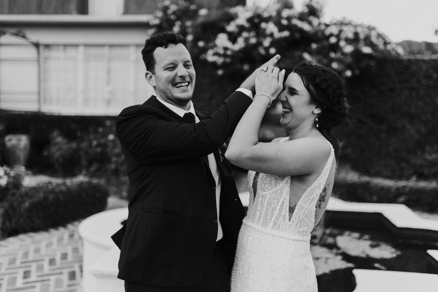 Groom playfully fixing brides hair while both laugh in candid black and white wedding photo.