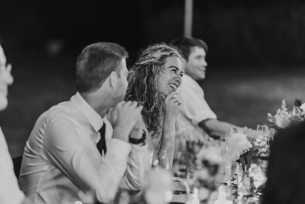 Bride with curly hair laughs joyfully at outdoor wedding reception dinner table.