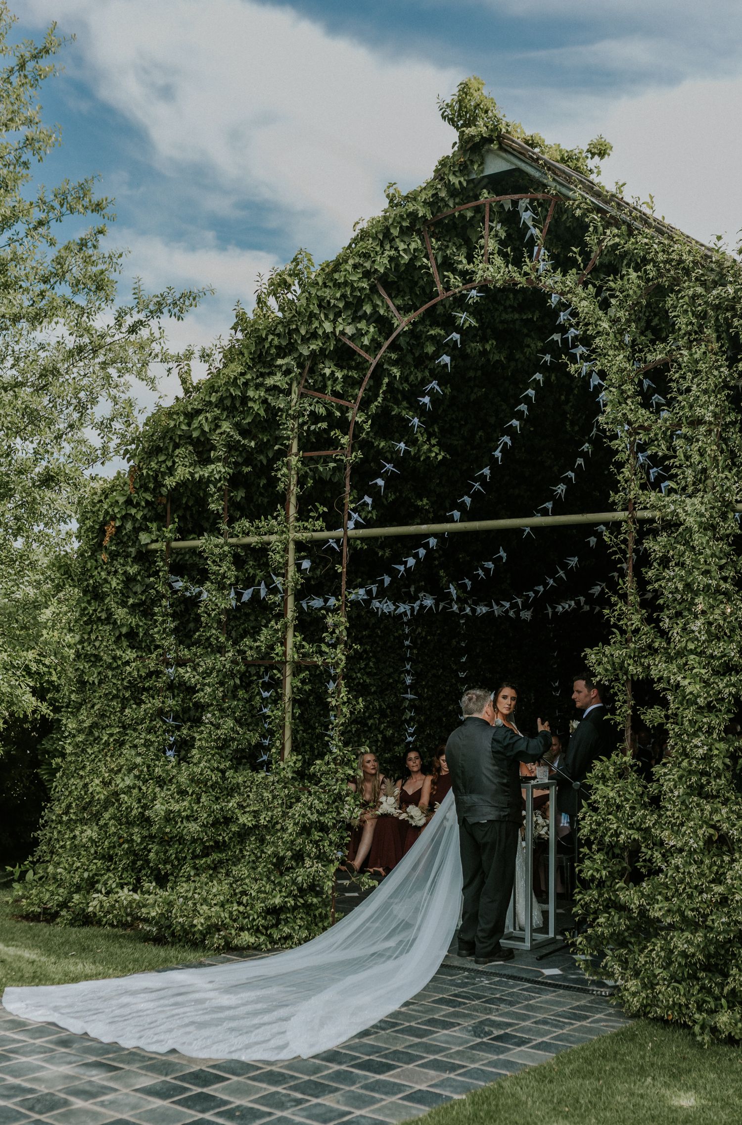 Bride and groom exchange vows beneath a stunning ivy-covered arch adorned with bunting decorations.