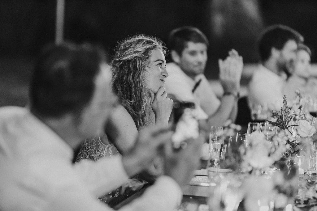 Bride applauds and smiles during wedding reception toasts at dinner table.