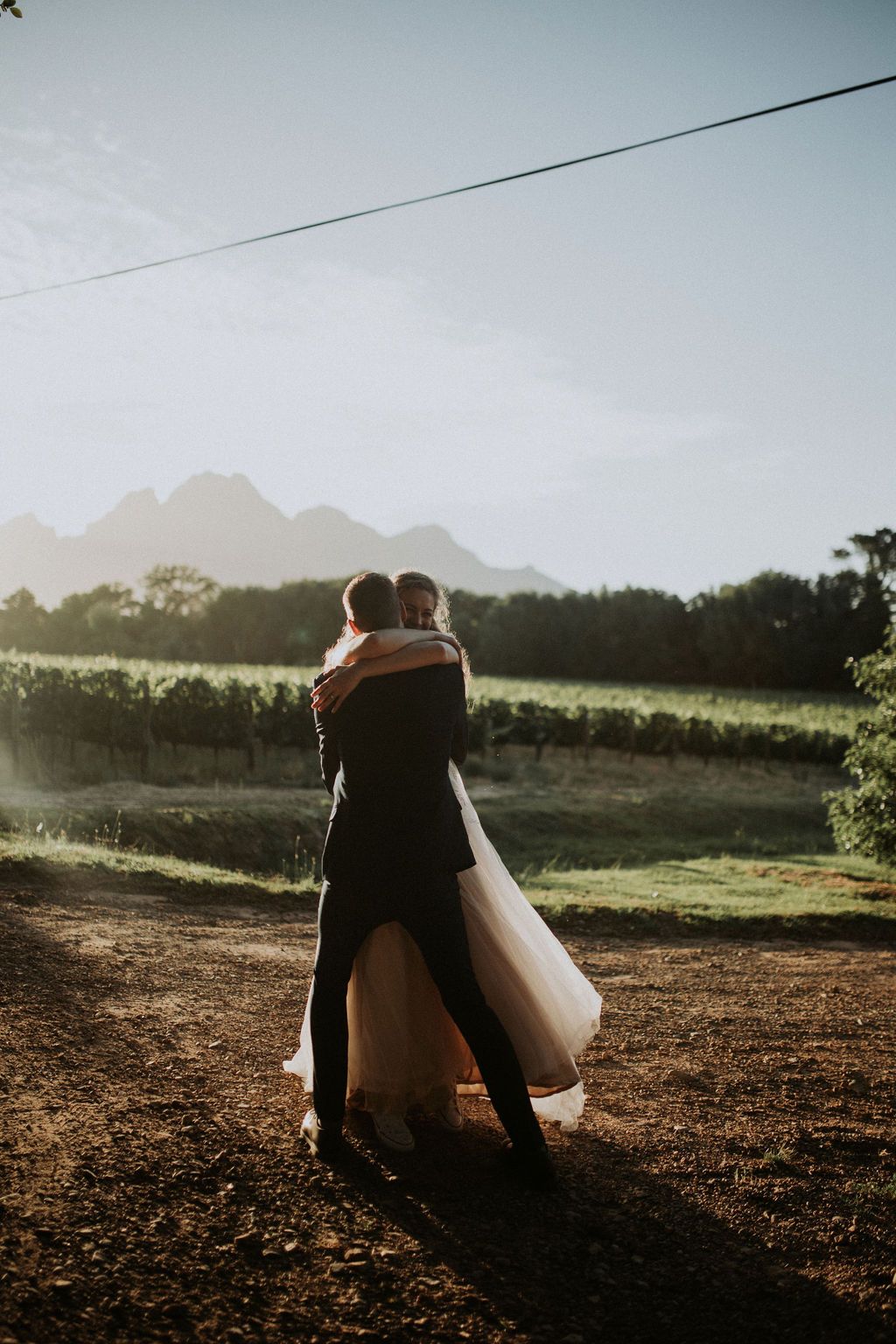Bride and groom embracing at sunset with mountain vineyard backdrop on their wedding day.
