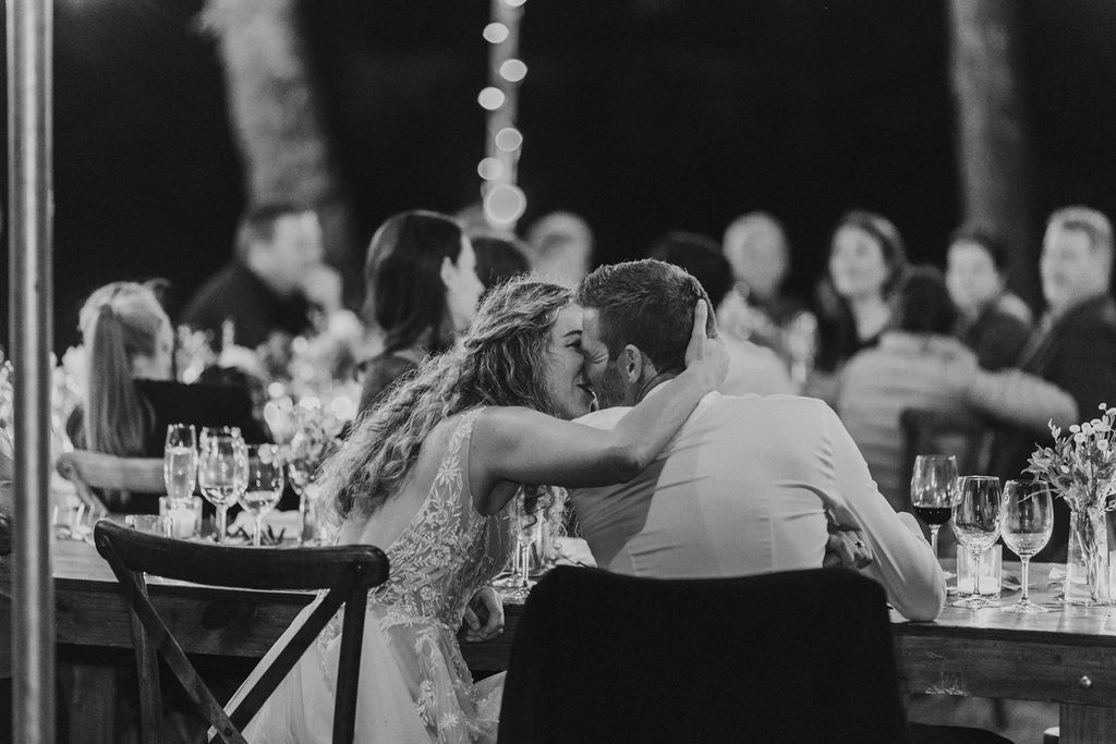 Bride and groom share romantic kiss at outdoor wedding reception table.