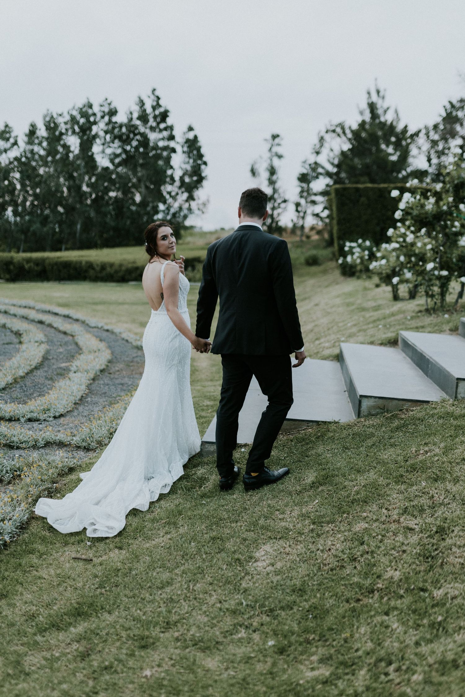 Bride and groom walk hand in hand through lush green wedding venue grounds with elegant white gown train trailing.
