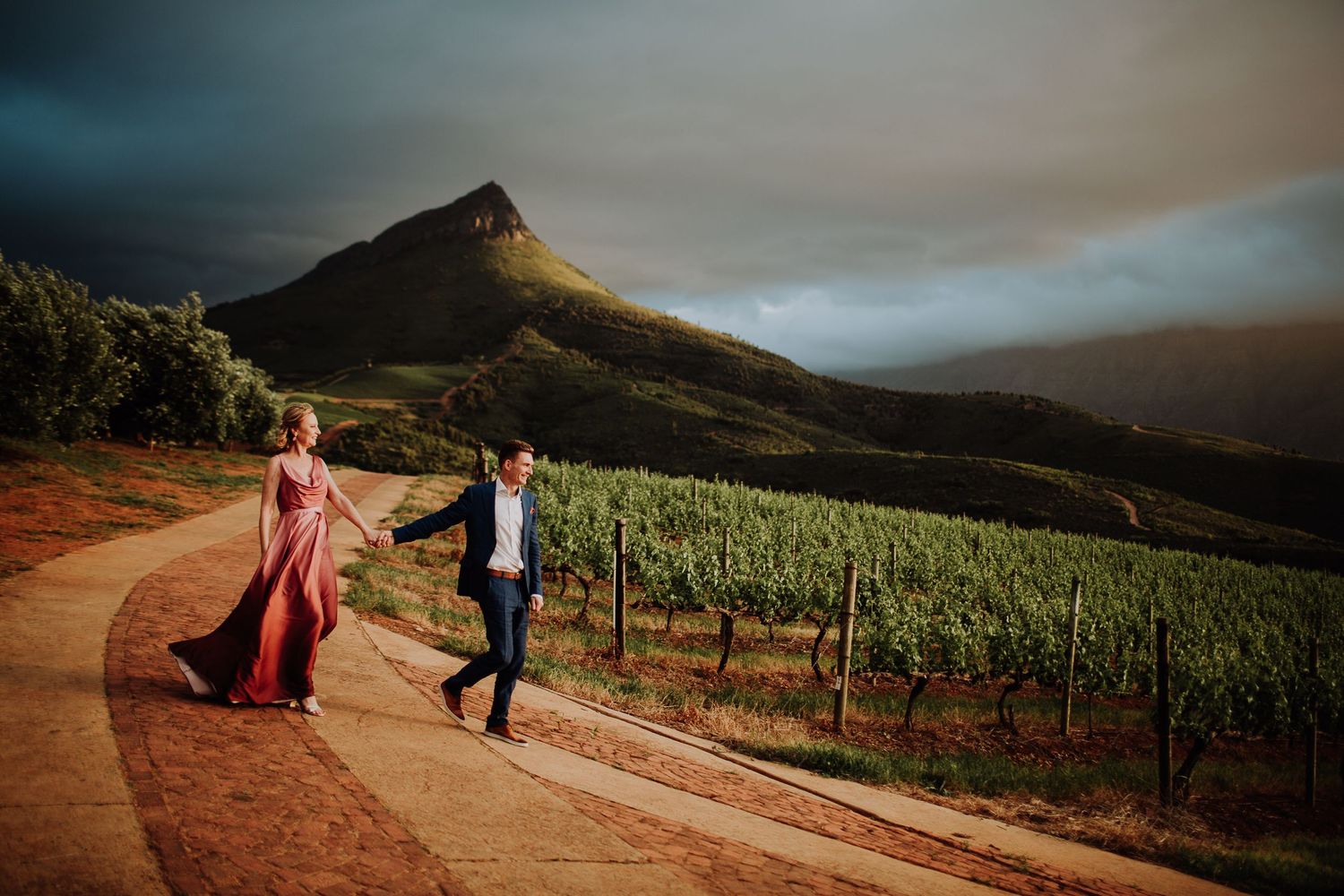 Romantic couple walking through a vineyard at sunset with moody storm clouds over a mountain peak.