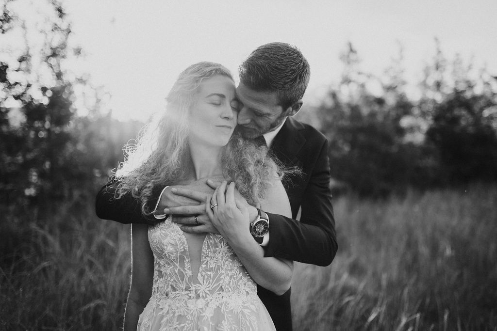 Black and white photo of groom embracing bride from behind in a sunlit field during golden hour.