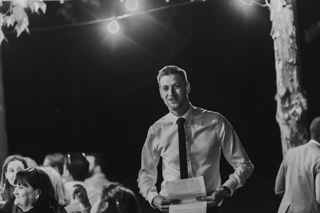 Best man holds speech notes while standing at outdoor wedding reception.