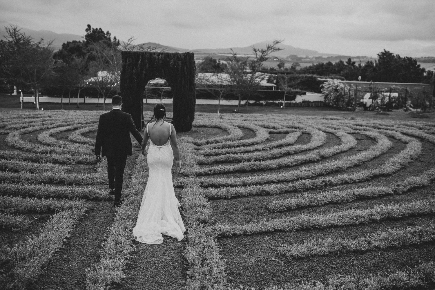 Black and white photo of bride and groom walking hand in hand through garden labyrinth.