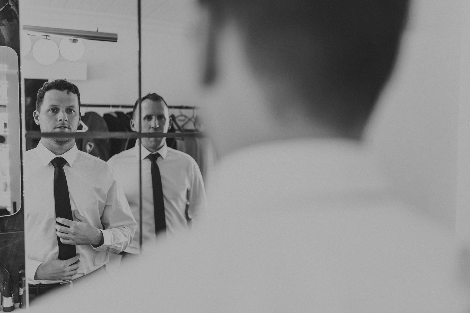 Groom adjusting his black tie while looking in a mirror in a black and white wedding photo.