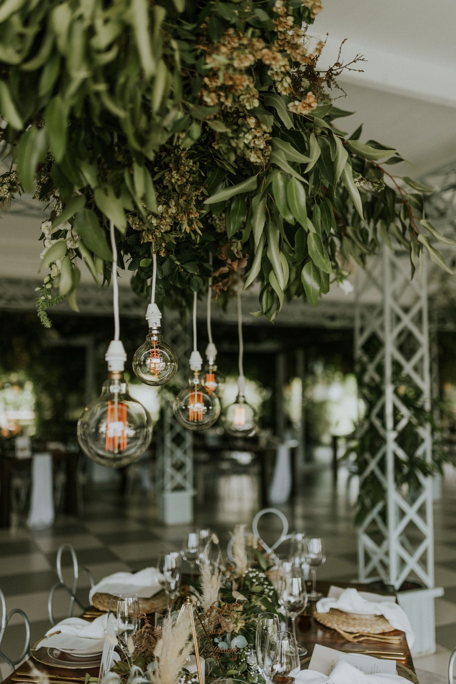 Rustic wedding tablescape with Edison bulb pendants hanging from a lush green floral installation.