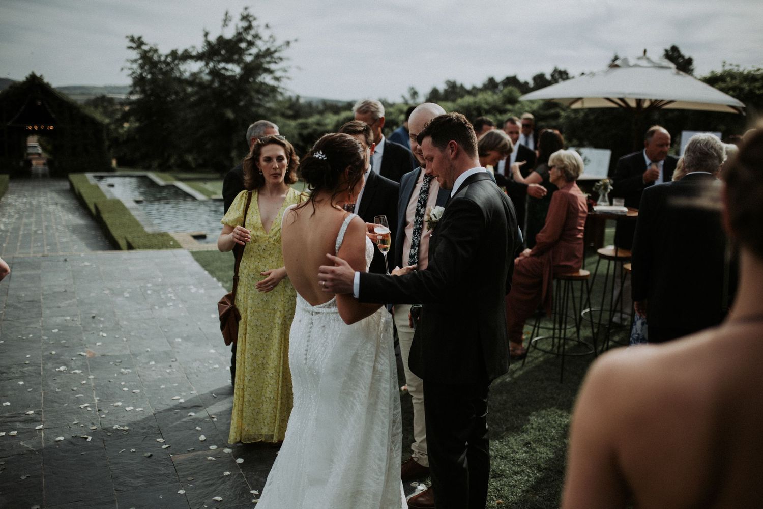 Bride and groom greeting wedding guests outdoors near a reflecting pool at an elegant garden venue.
