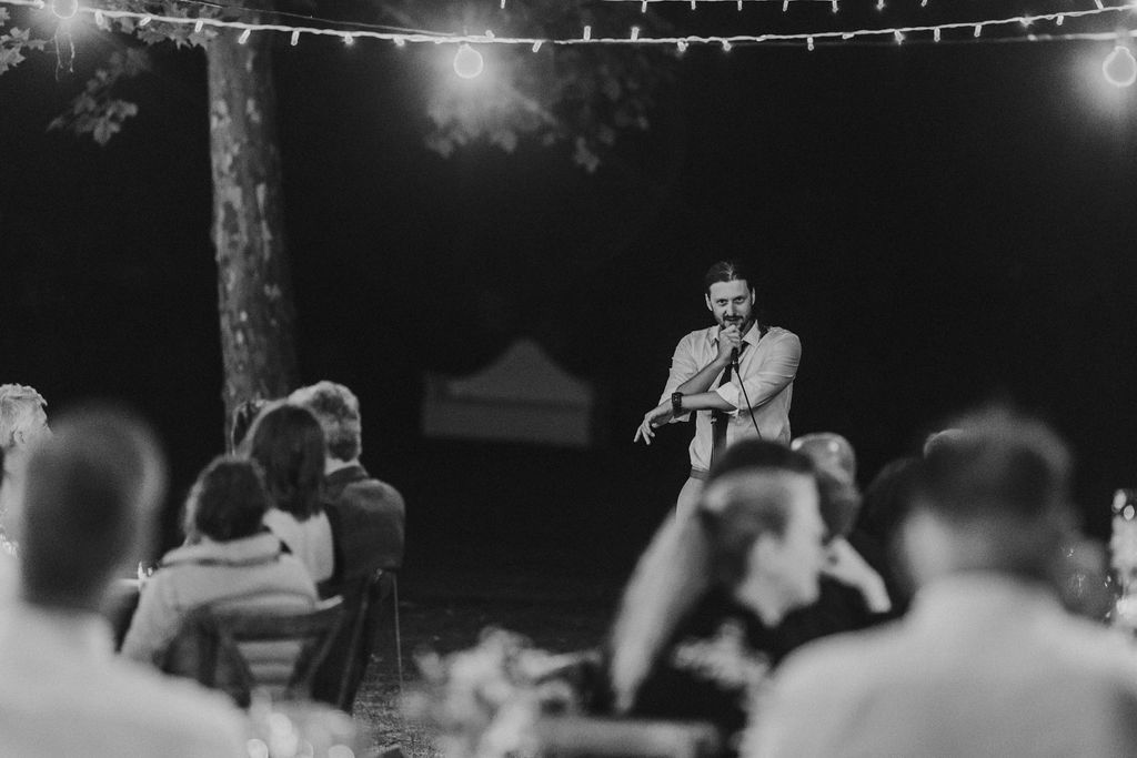 Speaker addresses seated guests at outdoor nighttime wedding reception with string lights above.