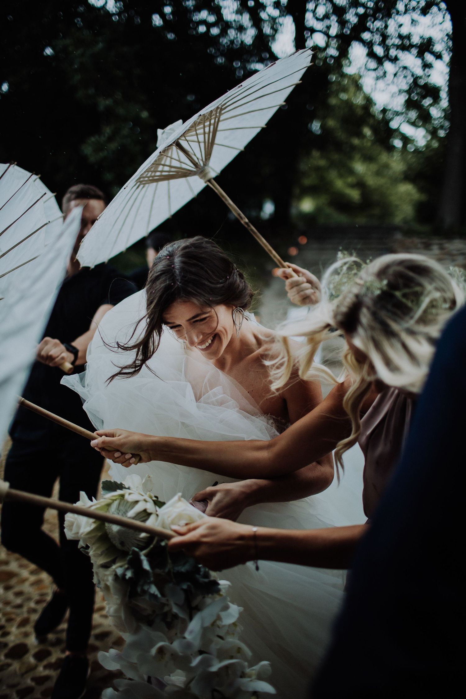 Radiant bride laughing while holding parasols with wedding party guests in a lush outdoor ceremony setting.