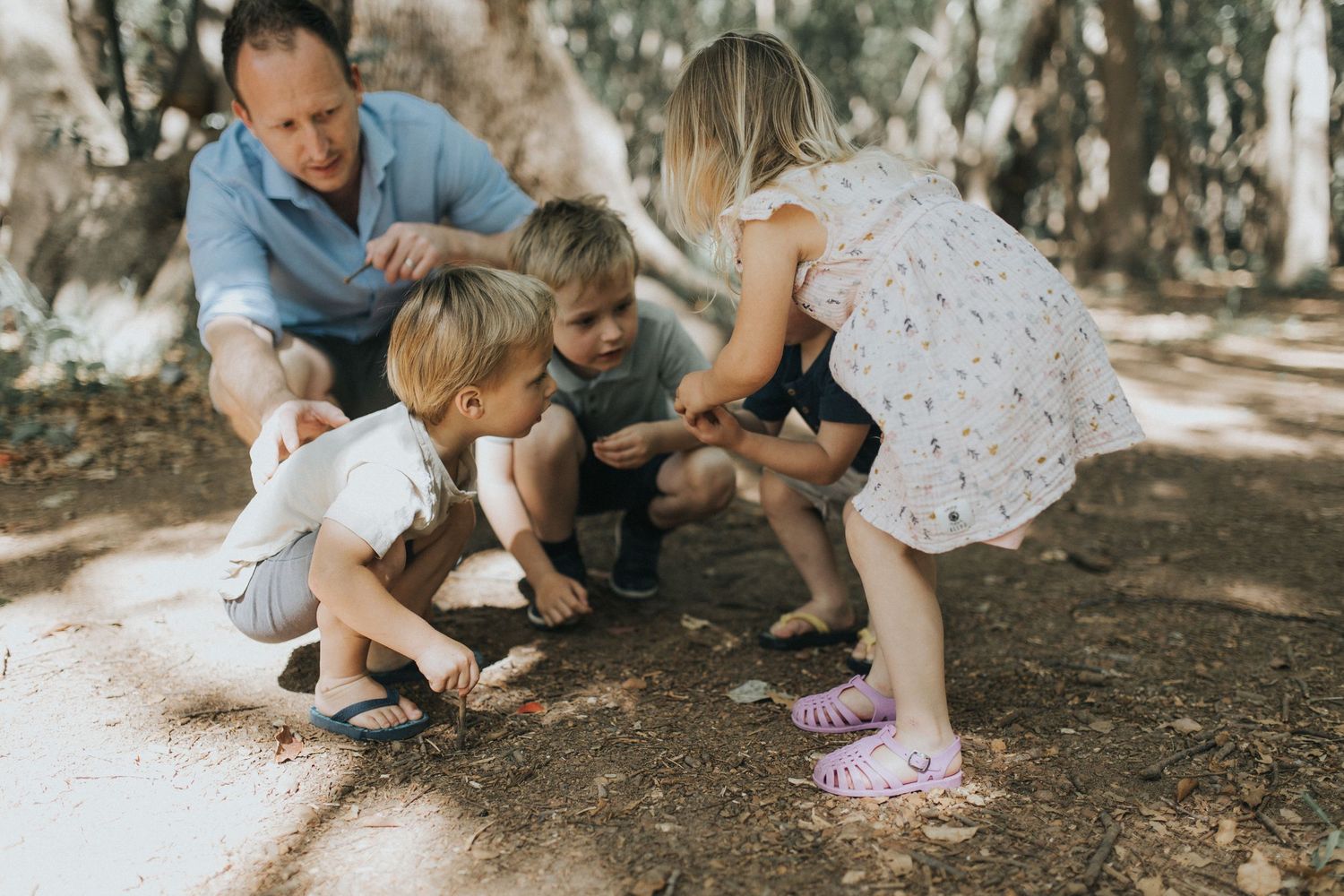 Father and three young children crouching together exploring nature on a woodland dirt path.