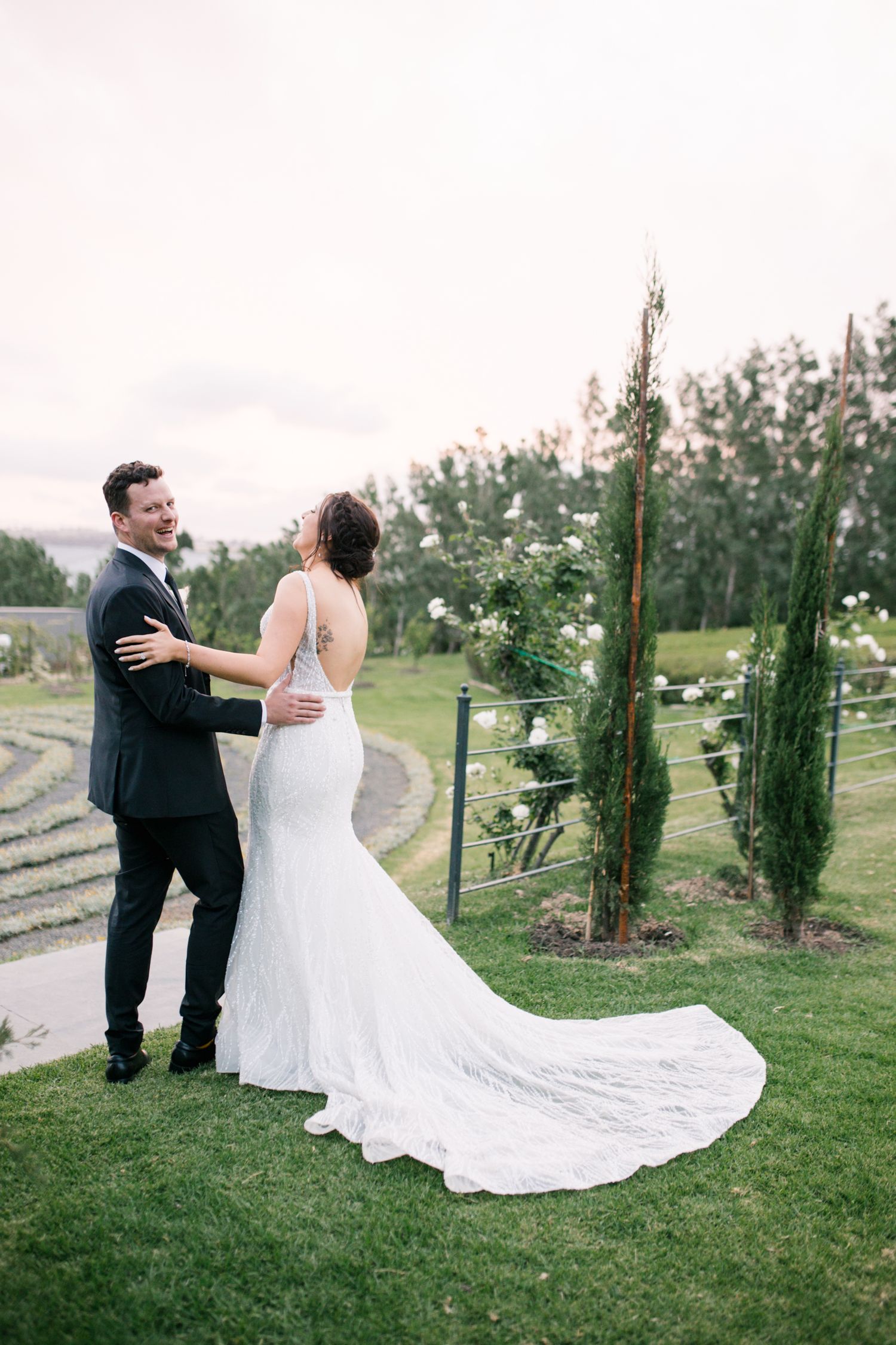 Bride and groom laughing together on lush green lawn with elegant wedding dress train spread out.