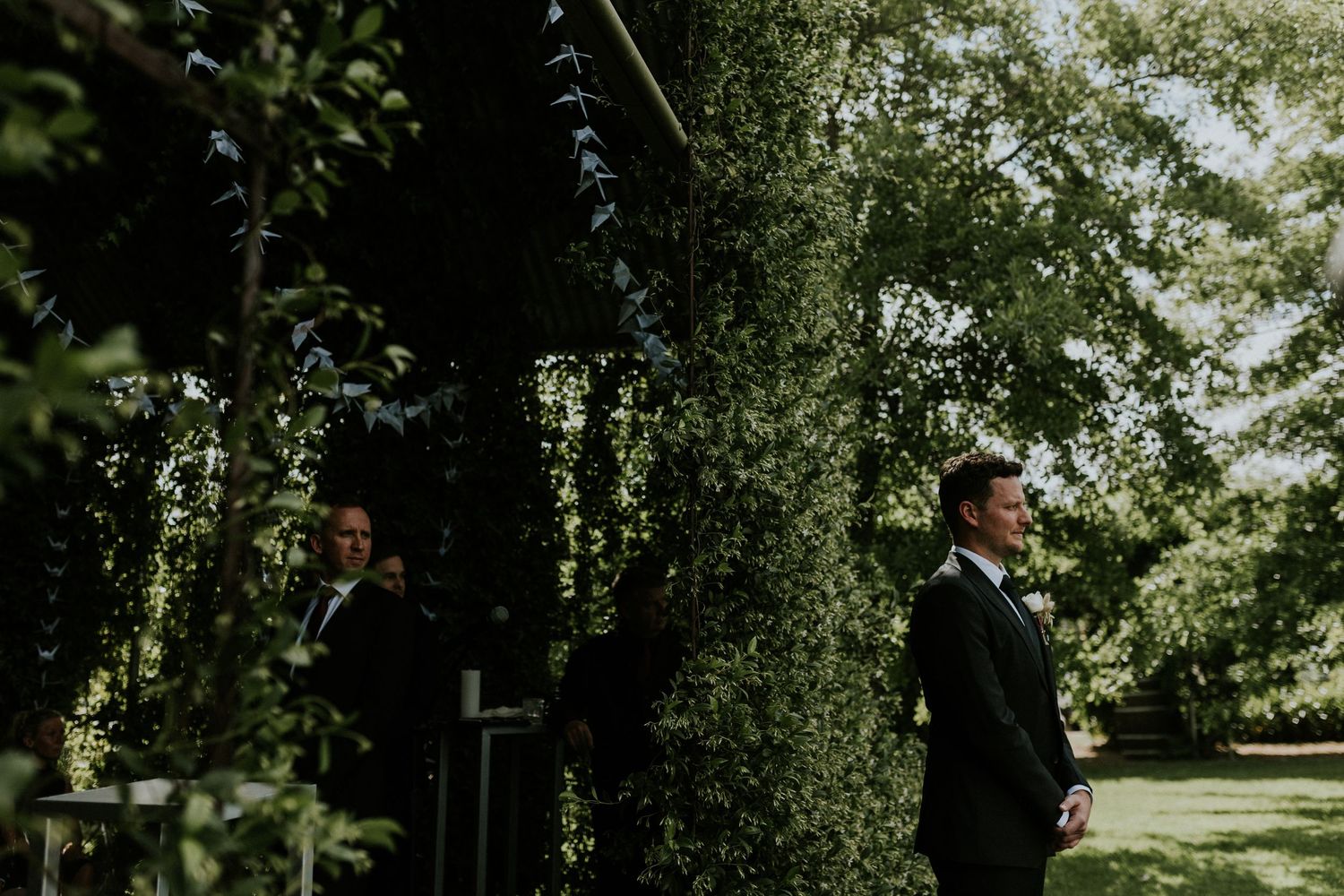 Groom in black suit standing among lush green trees before wedding ceremony.