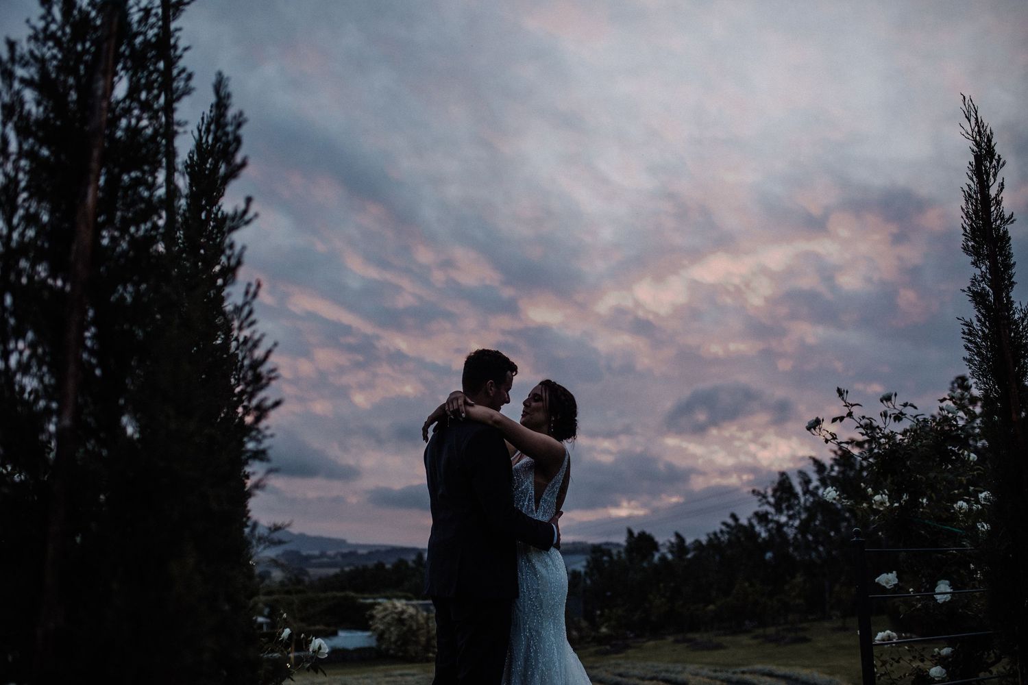 Silhouetted newlyweds embrace romantically beneath a dramatic pink and purple sunset sky.