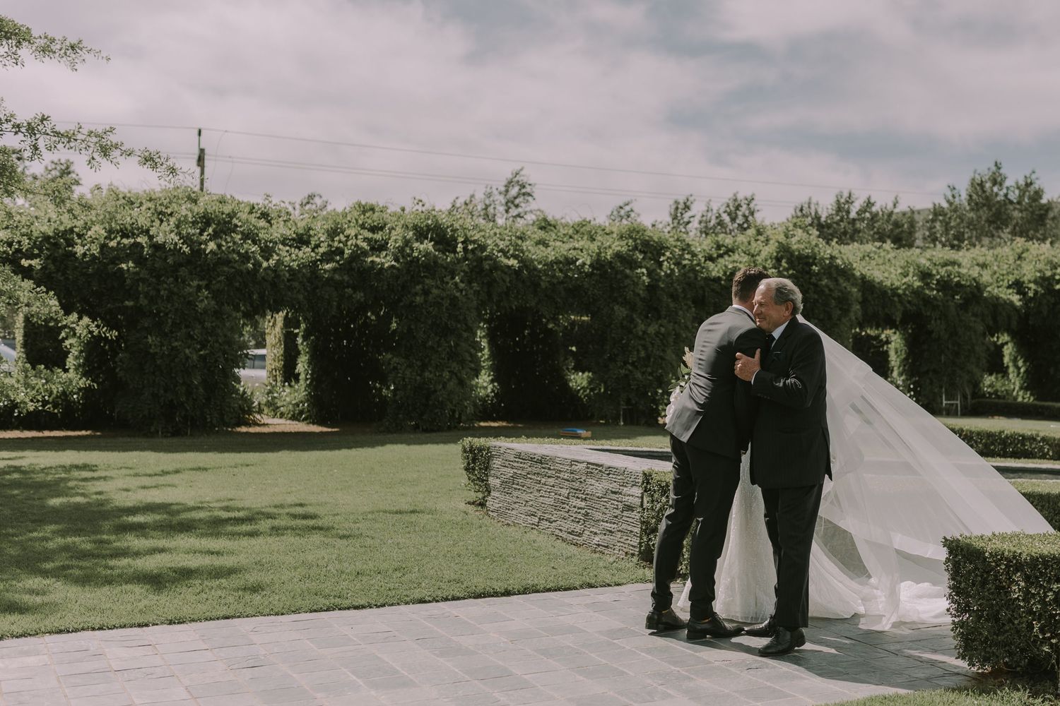 Tender moment as a bride in a flowing veil embraces her father in a lush green garden before the wedding ceremony.